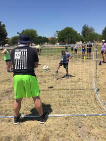 A coach calls for the ball from behind the soccer goal as an athlete attempts a shot.